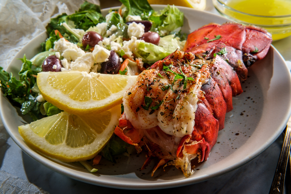 Grilled lobster tail on a plat with lemon slices, a salad and butter sauce.