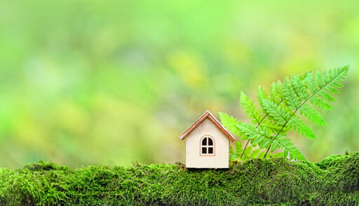 Miniature wooden house on a log next to a green leaf with a green forest in the background.
