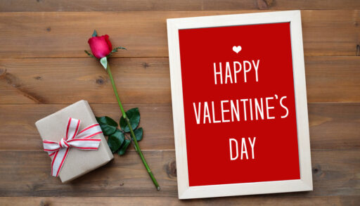 Red Valentine's Day sign on a wooden table with a wrapped present and a rose.