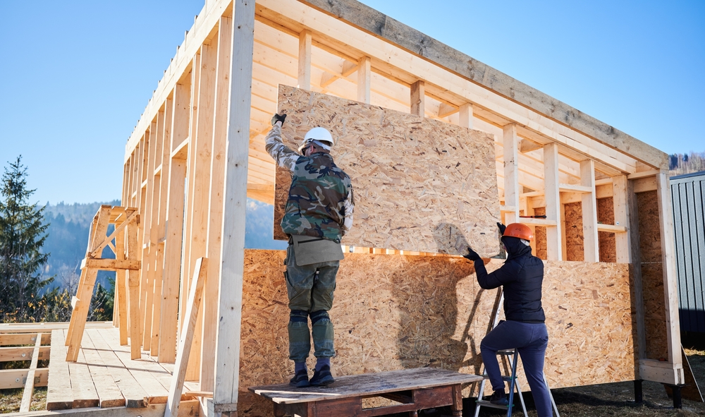 Two carpenters lifting a piece of plywood onto framing for a house.