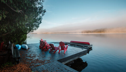 Red Muskoka chairs on a dock next to a still lake at sunset.