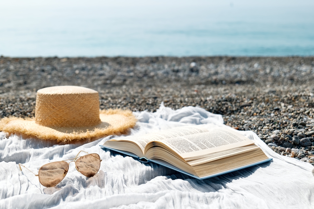 Hat, sunglasses and book laying open on a blanket next to water.