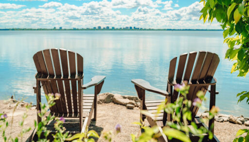 Two Muskoka chairs looking out over a blue lake with a city skyline on the horizon.