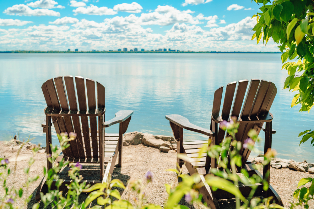 Two Muskoka chairs looking out over a blue lake with a city skyline on the horizon.