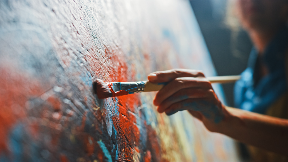 Person using a paint brush dipped in red paint to paint on a canvas.