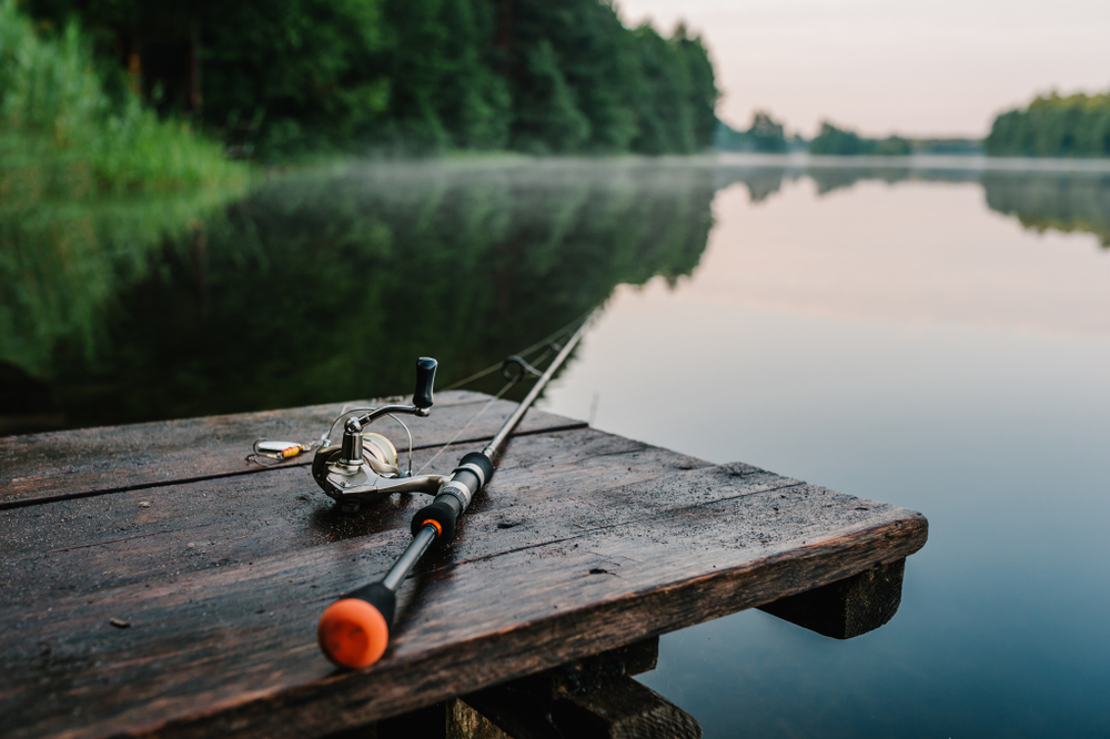 Fishing rod sitting on a wooden table overlooking a lake surrounded by trees.