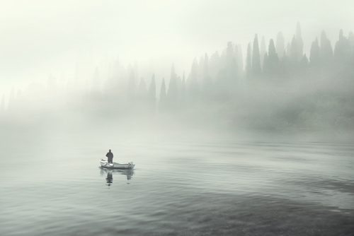 Man in a fishing boat on a fog-covered lake.