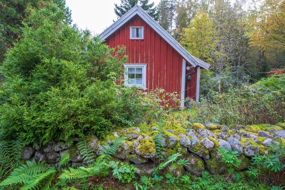 Small, red cottage next to a stone wall covered in flowers.