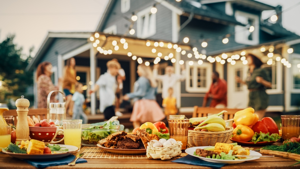 Outdoor table covered in food with a cottage and group of people in the background.