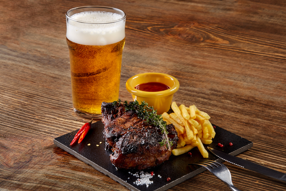 Glass of amber-coloured beer next to a plate with a steak and fries on it.
