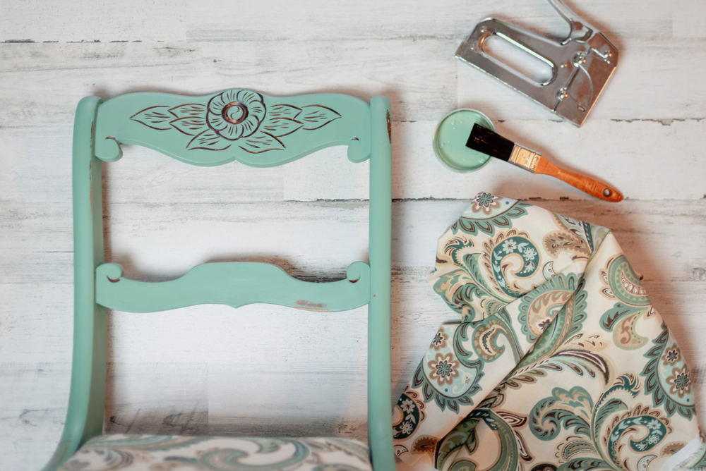 Teal-coloured antique chair next to a staple gun, paintbrush and fabric.