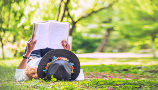 Woman wearing a grey hat reading a book in a field with trees.