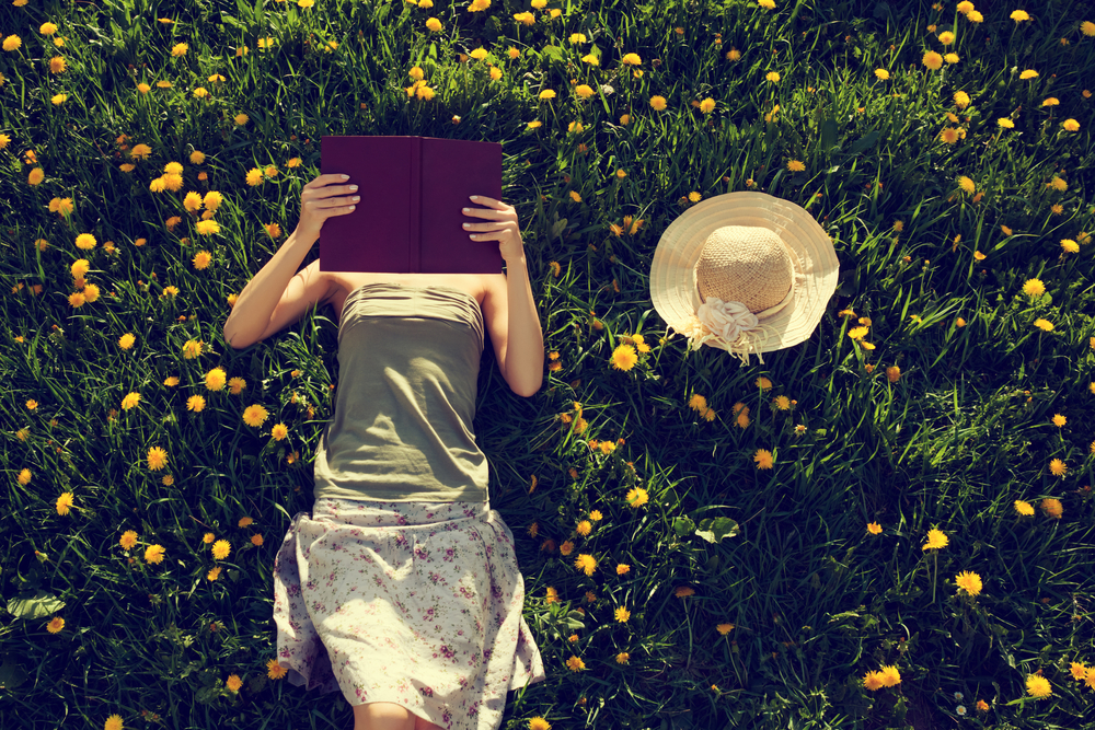 Girl laying in a field with yellow dandelions reading a book.