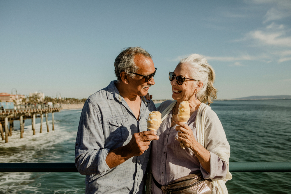 Older man and woman stand on a pier together eating ice cream.