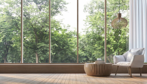Modern living room with wooden table and grey chair in front of large, panoramic windows looking into a forest.