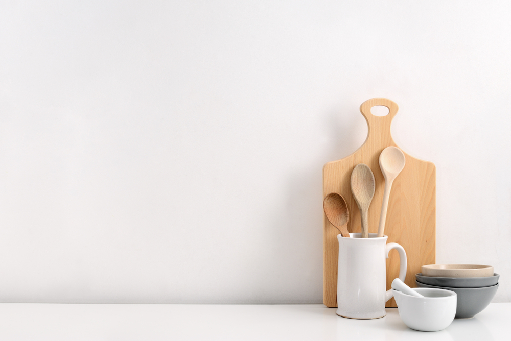 Wooden cutting board and white jar with wooden spoons on a white countertop.