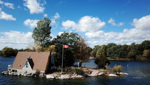 Small, grey cottage on an island in a lake next to a Canadian flag.