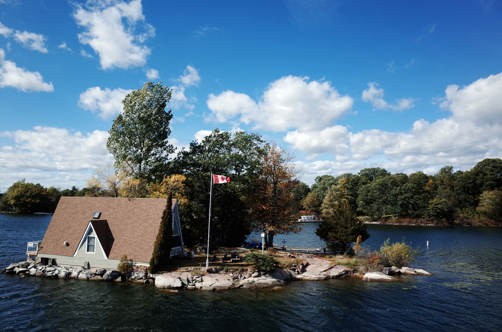Small, grey cottage on an island in a lake next to a Canadian flag.
