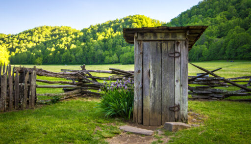 Wooden outhouse in a green farm field near a wooden fence.