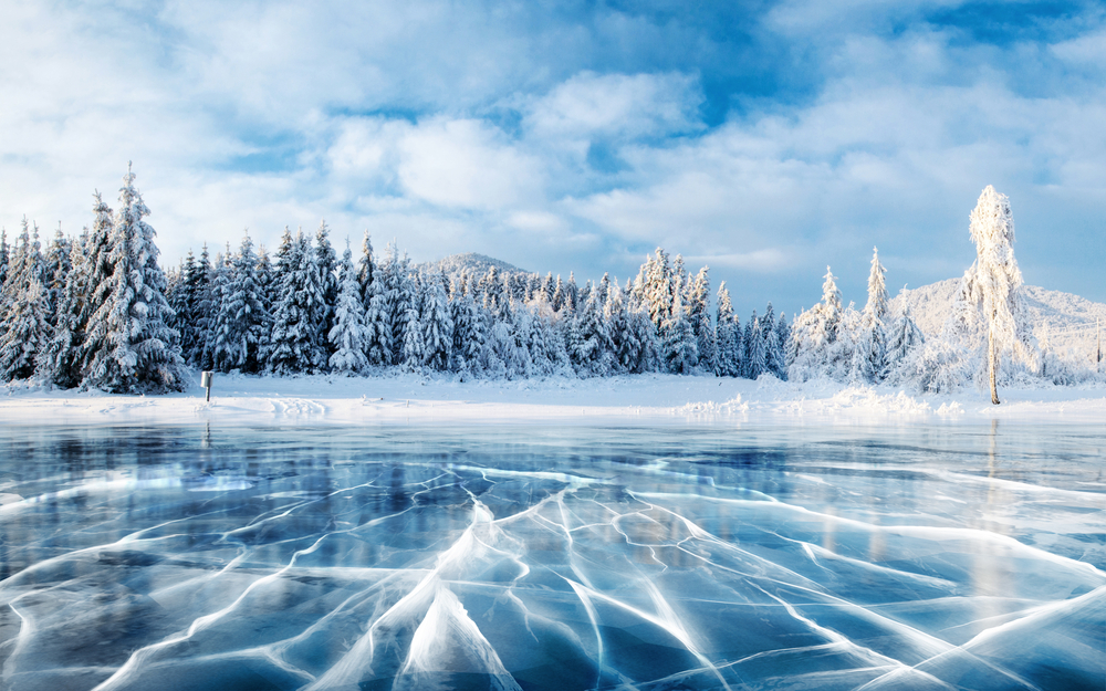 Cracks on the surface of a blue, frozen lake surrounded by snow-covered trees.