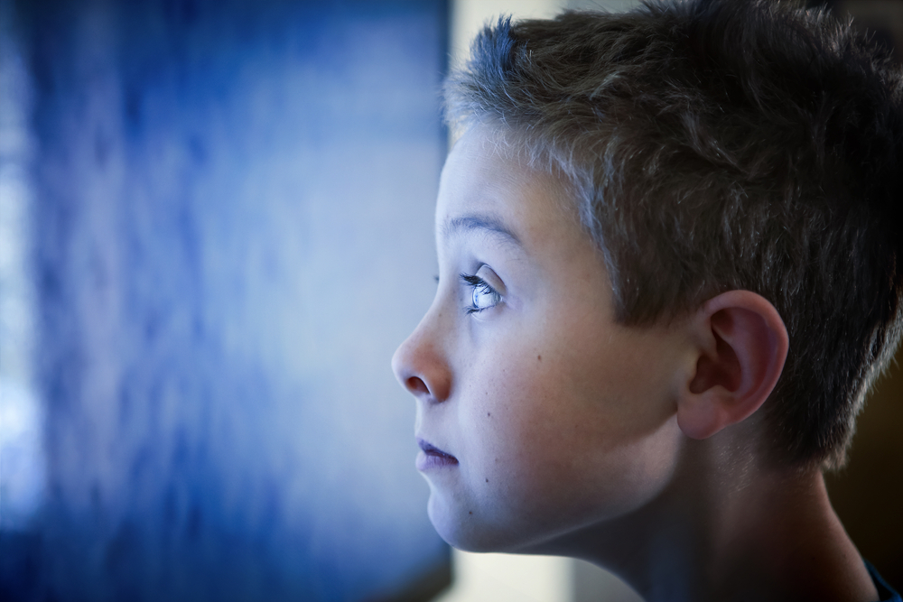 Close-up of a young boy staring at a blue television screen.