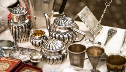 Antique silver teapot and tea cups on a table with other antique silverware.