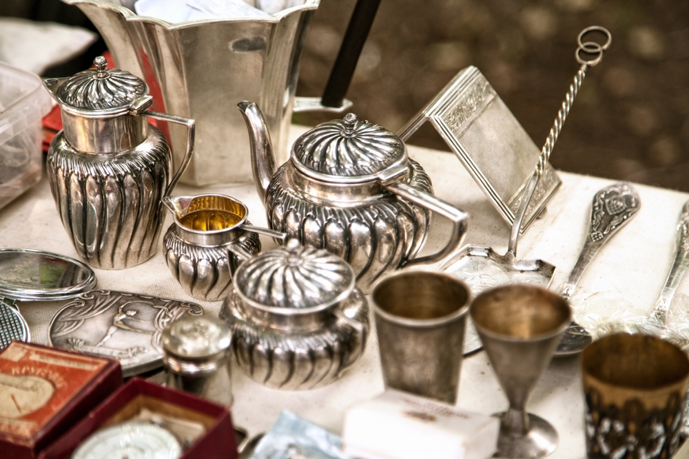 Antique silver teapot and tea cups on a table with other antique silverware.