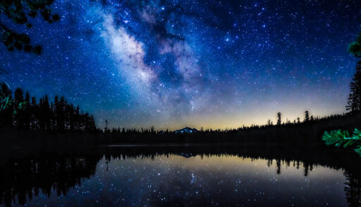 The Milky Way over a lake surrounded by silhouettes of trees.