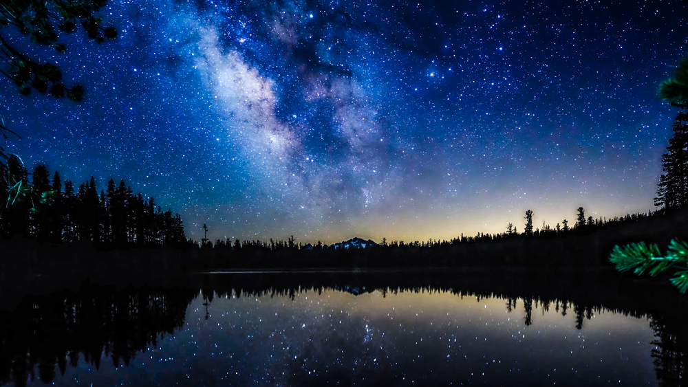 The Milky Way over a lake surrounded by silhouettes of trees.