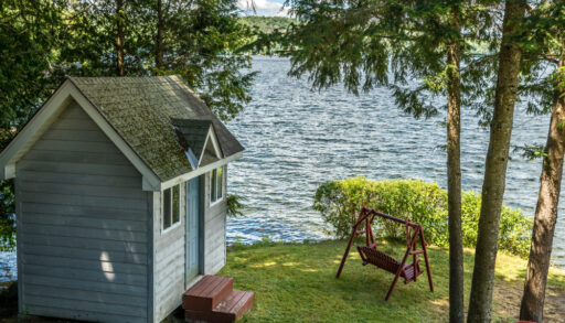 Small, light blue cottage bunkie next to a lake surrounded by trees.