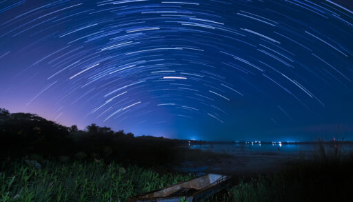 Star trail in a night sky over a hill with trees.