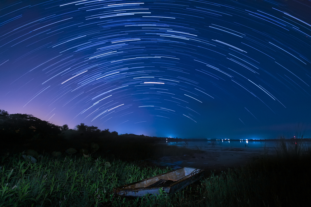 Star trail in a night sky over a hill with trees.