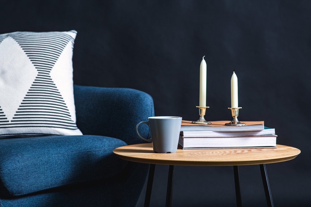 Blue armchair next to a wooden table with a stack of books, candles and a grey coffee cup.