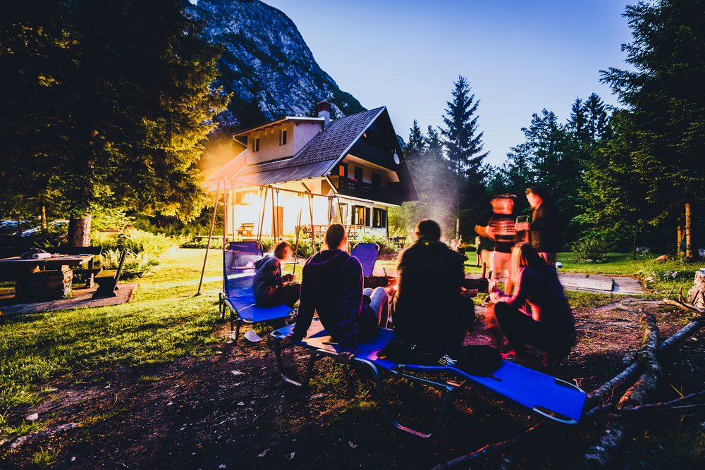 Group of people sitting around a campfire near a cottage in the evening.