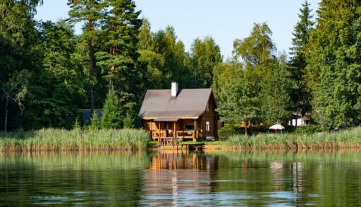 Small, wooden cabin next to a lake surrounded by green trees.