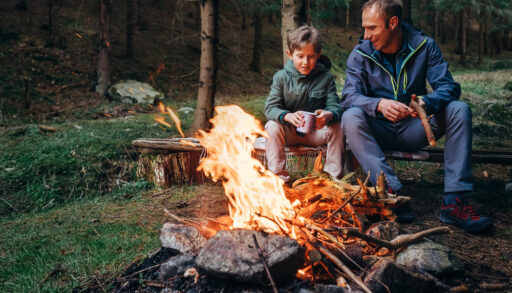 Father and son sitting on a wooden bench next to a campfire.