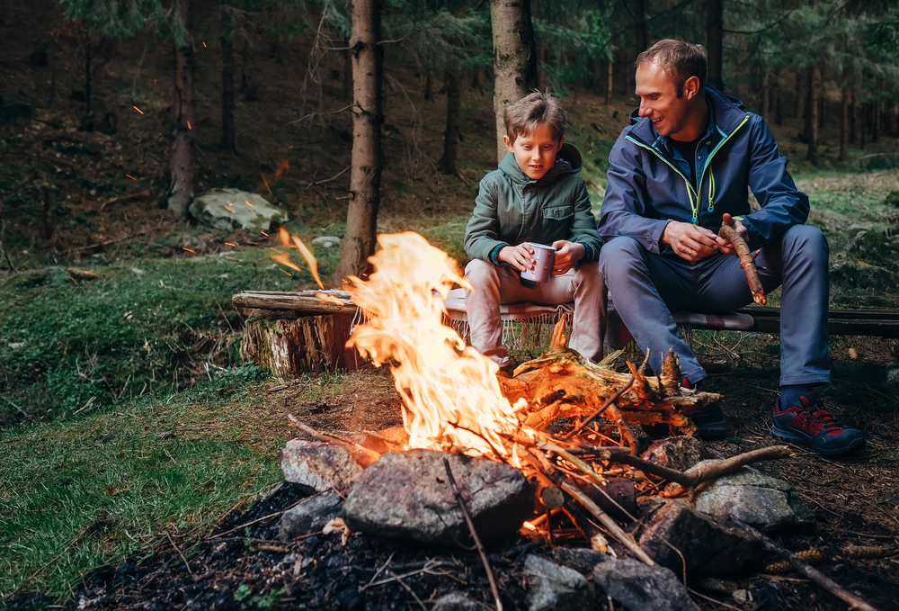 Father and son sitting on a wooden bench next to a campfire.