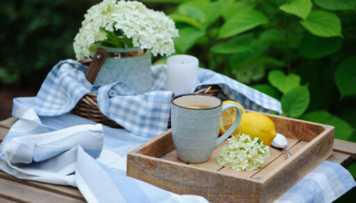 Wooden tray with a grey coffee mug on an outdoor table with a blue tablecloth and vase of flowers.