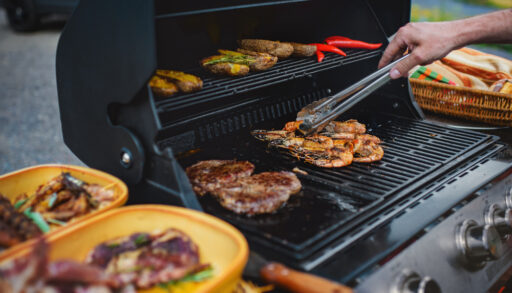 Person using metal tongs to flip various foods on a barbecue.