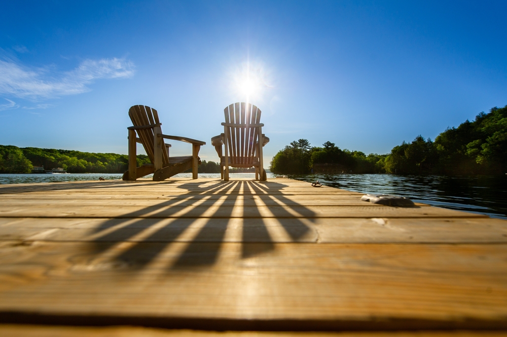 Low angle of sun shining through the back of a Muskoka chair on a dock next to a lake.