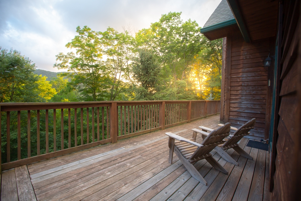 Two Muskoka chairs on a wooden deck overlooking the sun shining through a forest.