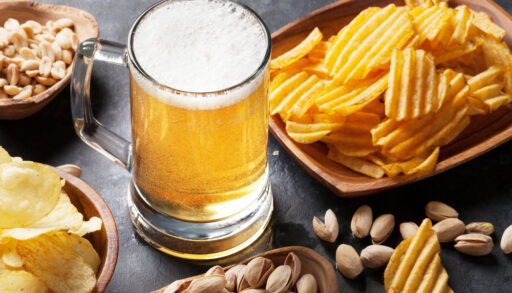 Glass of yellow-coloured beer on a table with chips and peanuts.
