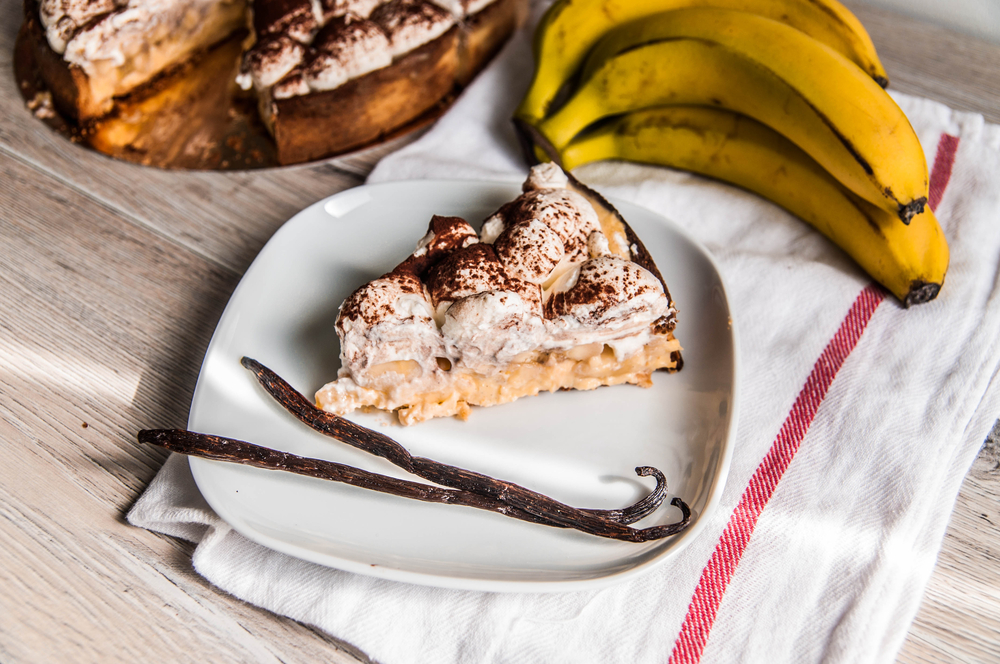 Piece of banofee pie on a white plate with two vanilla beans and a bunch of bananas.
