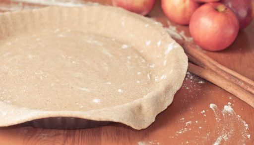 Close-up of an empty pie crust shell with cinnamon sticks and apples on the table next to it.