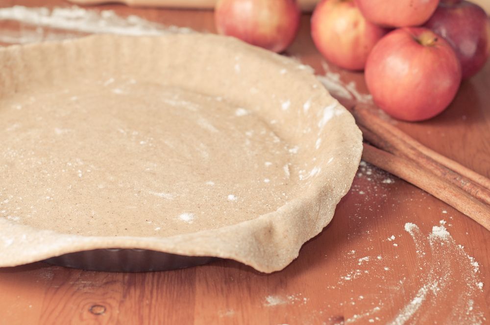 Close-up of an empty pie crust shell with cinnamon sticks and apples on the table next to it.