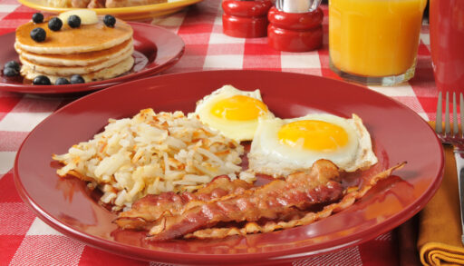 Plate of bacon and eggs on a red-checkered tablecloth with a plate of pancakes in the background.