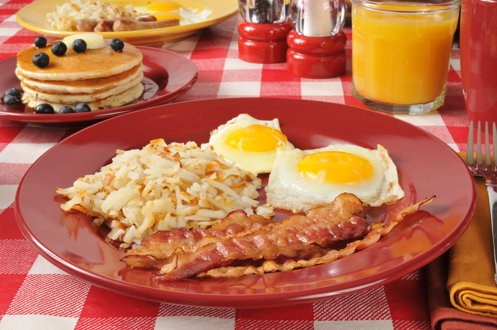 Plate of bacon and eggs on a red-checkered tablecloth with a plate of pancakes in the background.