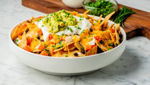 Bowl of nachos loaded with different toppings, next to a wooden cutting board with more toppings.