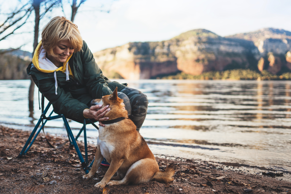 Young woman sitting next to a lake petting a dog.