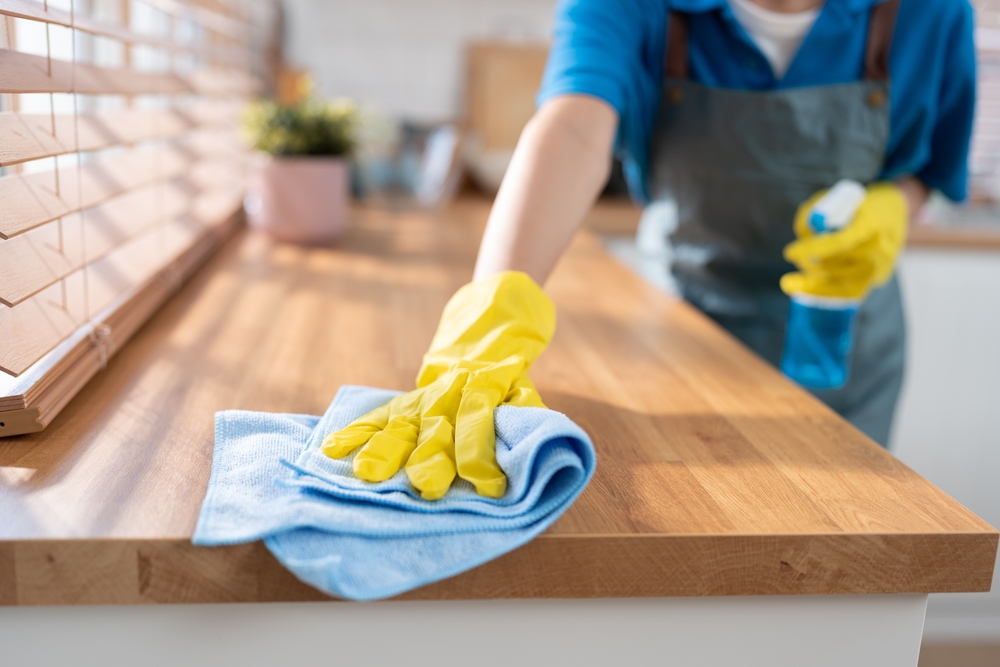 Close-up of a person wearing yellow rubber gloves cleaning a wooden countertop with a blue cloth.